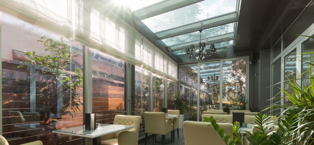 Modern terrace dining area featuring soft white Roman shades on large windows, filtering sunlight gently.