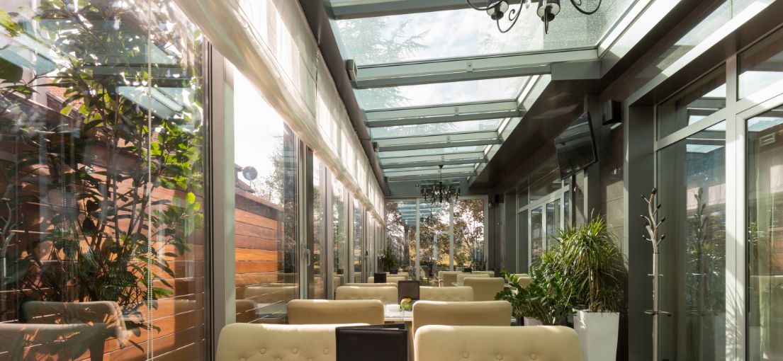 Interior view of restaurant terrace with ceiling-to-floor windows covered by light Roman shades, enhancing the airy ambiance.